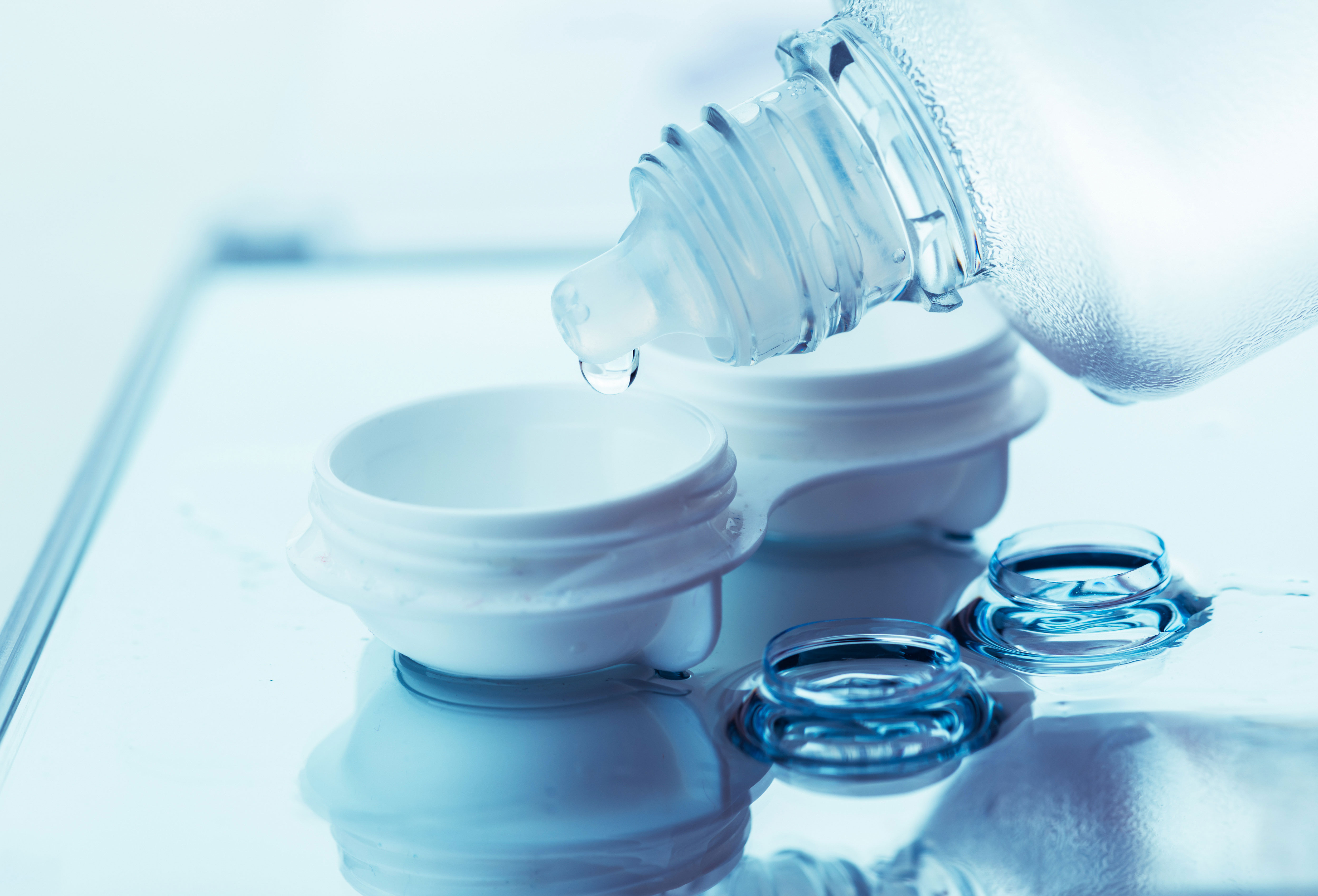 A bottle dripping solution into a case with contact lenses on a glass table.