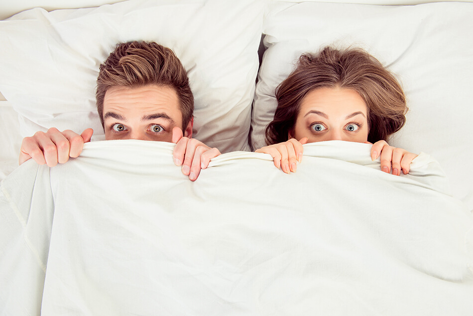 wide-eyed couple in bed with covers pulled up past their noses