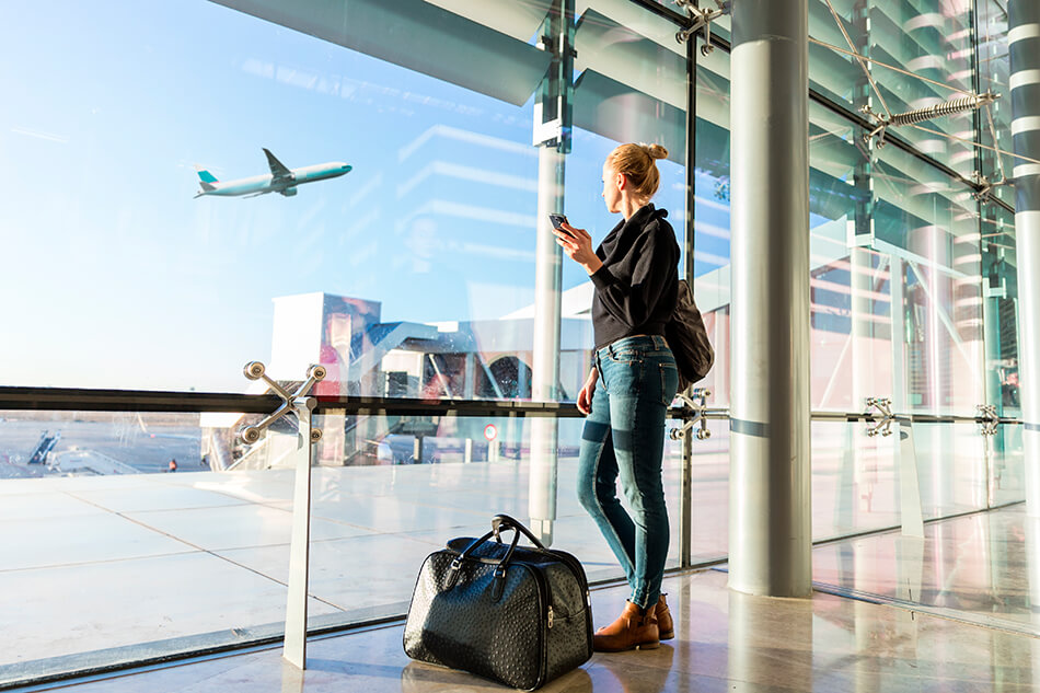 young woman standing at airport window next to her carry-on bag